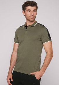 Olivgrünes Poloshirt mit schwarzen Schulterakzenten, weißem Kragenbesatz, kurzen Ärmeln und zwei Knöpfen am Hals. Weicher, glatter Stoff.