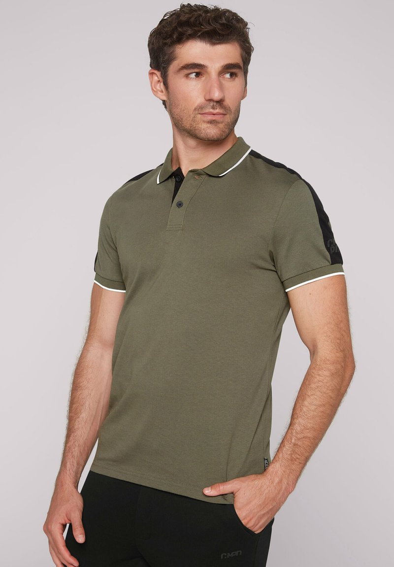 Olivgrünes Poloshirt mit schwarzen Schulterakzenten, weißem Kragenbesatz, kurzen Ärmeln und zwei Knöpfen am Hals. Weicher, glatter Stoff.