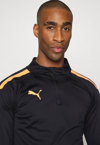 Schwarzes sportliches Oberteil mit halbem Reißverschluss, orangefarbenen Schulterstreifen und Puma-Logo auf der Brust. Glatte, leichte Stofftextur.