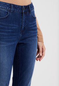 Primer plano de una persona que lleva jeans de mezclilla azul oscuro y un suéter color marrón claro, con una mano descansando relajada a su lado.