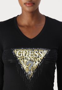 Kvinna som bär svart V-ringad skjorta med triangel-Guess-logga i leopardtryck och strassdetaljer på bröstet.