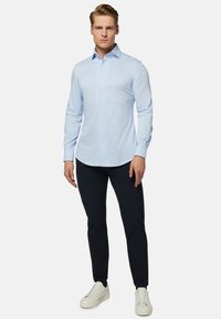 Chemise bleu clair à manches longues, en tissu lisse, avec des boutons blancs, assortie à un pantalon foncé et des baskets blanches. Design simple et structuré.