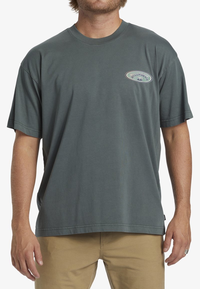 Homme portant un t-shirt Billabong vert foncé ample et un pantalon beige, se tenant avec les mains détendues sur les côtés.