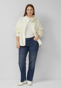 Femme portant une veste polaire blanche, un col roulé à côtes blanc, un jean bleu foncé, des baskets blanches, debout avec une main dans la poche contre un mur gris.