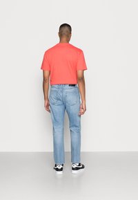 Homem de costas, vestido com uma camisa coral de manga curta, jeans azul claro e ténis preto e branco da Nike, contra um fundo simples.