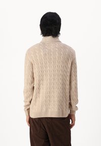 Pull en tricot beige avec un col haut, des poignets et un ourlet côtelés. Le motif texturé ajoute de la profondeur ; matériau doux pour le confort.