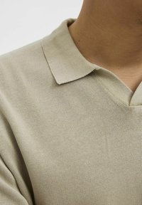 Beige gebreide polo met een gestructureerde oppervlakte, voorzien van een v-hals en een gestructureerde kraag. Minimalistisch ontwerp zonder zichtbare patronen of accenten.