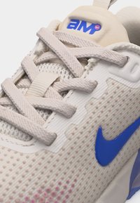 Κοντινή λήψη μπεζ αθλητικού παπουτσιού με λευκά κορδόνια, μπλε λογότυπο Nike στο πλάι και επάνω μέρος από δίχτυ με μικρές ανυψωμένες τελείες.