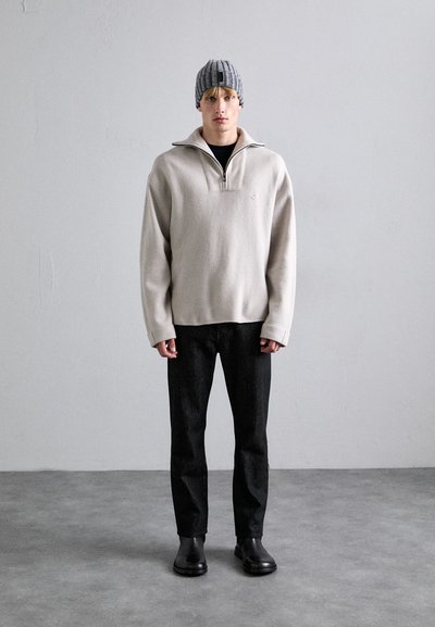 Beige fleece pullover with a half-zip collar, dark jeans, and black boots. The model wears a gray ribbed knit beanie. Simple design, textured fabric.