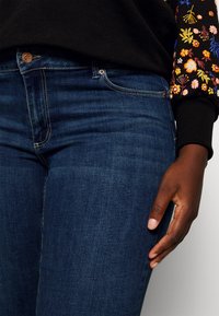 Dark blue denim jeans with classic five-pocket design, featuring a copper button and stitching. Hand rests lightly on the thigh.