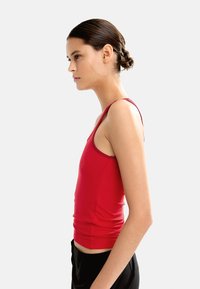 Czerwony dopasowany top na ramiączkach z cienkimi paskami, o gładkiej fakturze i wysokim dekolcie, noszony z czarnymi spodniami. Widok z boku.