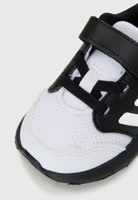 Weißer Sneaker mit schwarzen synthetischen Akzenten, ausgestattet mit einem Obermaterial aus Mesh, schwarzen Schnürsenkeln und einem Klettverschluss für einen sicheren Sitz. Strukturierte Laufsohle.