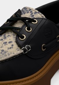 Chaussure bateau en cuir noir avec languette imprimée léopard, lacets noirs, œillets en métal et semelle en caoutchouc marron clair, avec logo embossé sur le côté.