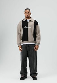 Grijs-zwart kleurblokkend jack met rits en logo, met een relaxte pasvorm en geribde manchetten, gecombineerd met losse donkere jeans en zwarte schoenen.