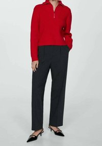 Persona che indossa un maglione rosso con zip, pantaloni dritti grigio scuro e décolleté nere con punta appuntita, in piedi con una mano in tasca.