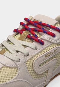Αθλητικό παπούτσι με κρέμ διχτυωτό ύφασμα και σουέτ, χρυσές λεπτομέρειες, κορδόνια σε κόκκινο και μπλε που επικαλύπτουν λευκά κορδόνια, και λογότυπο "REPLAY" στην πλευρική επιφάνεια.