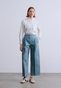 Lichtblauwe broek met brede pijpen, plooien en een hoge tailleband, gecombineerd met een wit overhemd met knopen. Kenmerkt zich door strakke lijnen en een eenvoudig ontwerp.