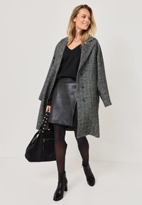 Manteau en tweed pied-de-poule gris avec un large col, associé à un haut noir et une jupe en cuir. Bottines noires, collants et un grand sac noir à clous.
