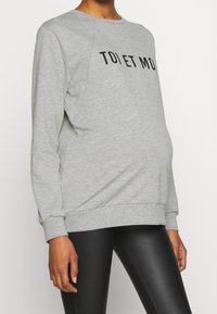 Sweat-shirt gris à finition texturée, arborant un slogan imprimé en noir. Manches longues avec poignets côtelés. Associé à des leggings noirs.