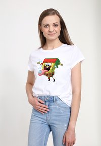 Hvid t-shirt med et farverigt SpongeBob SquarePants grafik, iført en grøn hat, røde ternede shorts og legende detaljer som bobler.
