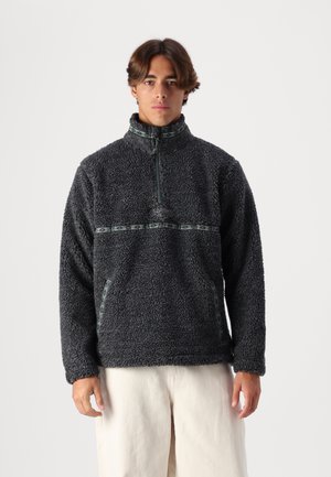 Black fleece pullover featuring a high collar, half-zip front, embroidered logo, and patterned green accent stripe across the chest.