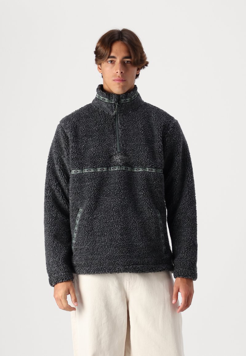 Black fleece pullover featuring a high collar, half-zip front, embroidered logo, and patterned green accent stripe across the chest.