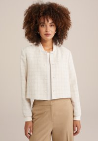 Blouson court en tissu crème texturé avec un col rond, des boutons-pression et des poignets côtelés. Associé à un pantalon beige.