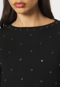 Femme portant un haut noir orné de petits clous circulaires dorés dispersés sur le tissu, gros plan sur le cou et le haut de la poitrine.