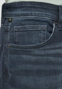 Des jeans en denim bleu foncé présentent un design classique avec une poche arrière, un léger décoloré, et une petite étiquette noire sur la ceinture.