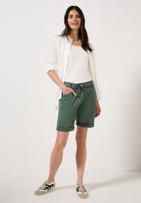 Groene shorts met een trekkoord in de taille en opgerolde mouwen, gecombineerd met een wit tanktop en een lichtwit overhemd. Casual schoeisel met accenten.