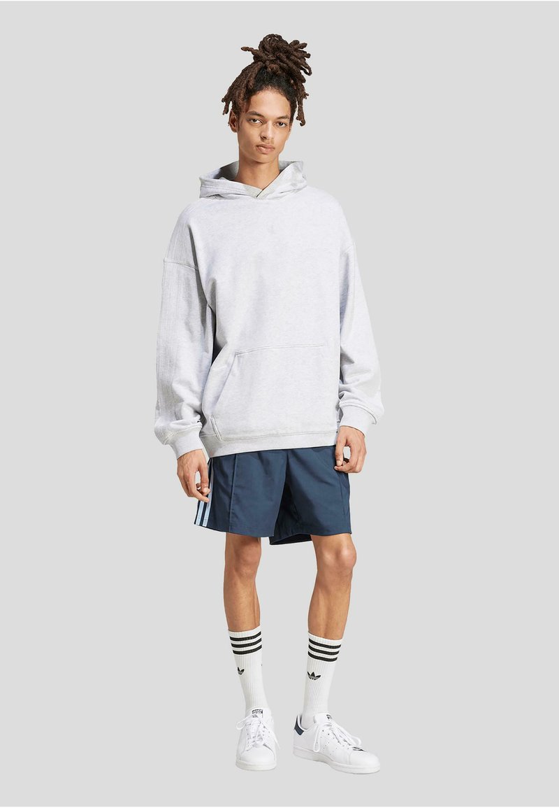 Haut à capuche oversize gris clair avec une poche frontale, associé à un short bleu marine avec des rayures blanches sur les côtés. Des baskets blanches et des chaussettes rayées complètent le look.