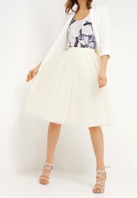 White tulle midi skirt with a fitted waistband, paired with a floral patterned top and a white blazer; model wearing beige strappy heels.
