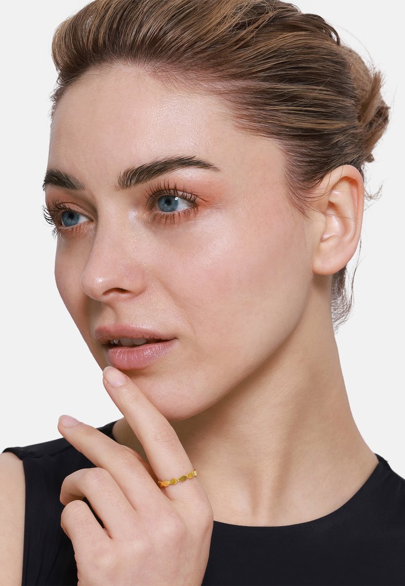 Bague en or ornée de petites pierres précieuses rondes et ovales, avec un design de bande délicat. Le modèle pose un doigt sur son menton, mettant en valeur la bague et sa main.