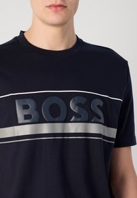 Tamsiai mėlynas medvilninis marškinėlis su ryškiu iškilniu "BOSS" logotipu, sidabrinėmis horizontaliomis juostelėmis ir klasikiniu apvaliu kaklu.
