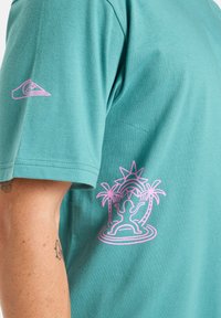 Tyrkysové bavlněné tričko s kulatým výstřihem. Na levém rukávu má růžovou grafiku postavy a palem. Hladký materiál, krátké rukávy.