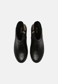 Botas de tobillo de piel negra con un upper texturizado, detalle de cadena dorada y diseño de punta redonda lisa. Nombre de la marca impreso visiblemente en el interior.