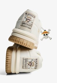Sneaker bianca con suola in gomma, pelle testurizzata e una toppa "Wanted" con un'illustrazione di un personaggio. Include un ciondolo a tema pirata.