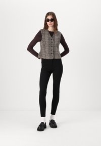 Beige un brūns tweed veste ar pogu priekšpusi, valkāta pār tumši brūnu garo piedurkņu topu un melniem legingiem, papildināta ar melnajām kurpēm.