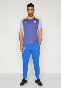 Treningsantrekk med en lilla Nike-skjorte med korte ermer og gule detaljer, kombinert med blå bukser og svart fottøy.