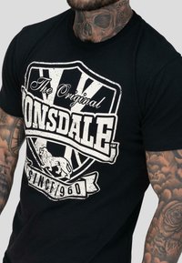 Mies, jolla on tatuoidut käsivarret ja kaula, pukeutuneena mustaan t-paitaan, jossa on valkoinen "Lonsdale Since 1960" -logo, jossa on leijonavaakuna.