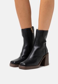 Bottes à cheville en cuir noir avec un bout carré, un détail de fermeture éclair dorée et un talon épais en bois. Texture lisse avec un design élégant.