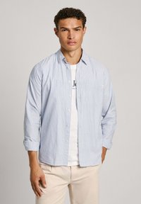 Camisa de botones de rayas azul claro y blanca con mangas largas, que cuenta con un bolsillo en el pecho y se lleva sobre una camiseta blanca, combinada con pantalones beige.