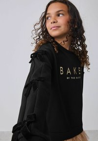 Musta huppari, jossa on kultaisella kirjotettu "BAKER" ja "by TED BAKER", featuring samettiset nauhat hihoissa ja ribbaattu helma.