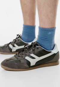 Klassiske atletiske sneakers med en overdel i brunt ruskind og læder, hvide accenter, brune snørebånd og en gummirubsål. Blå ribbede sokker synlige.