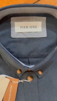 Camisa azul escura de colarinho com um design de botão. Apresenta botões de madeira e um tecido acolchoado e texturizado. Rótulo da marca: "PIER-ONE."