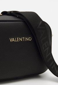 Svart lærveske med strukturert overflate, gull "VALENTINO"-logo og en svart stropp med hevet tekstdesign. Kompakt rektangulær form.