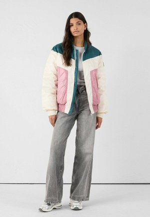 Donna in piedi che indossa un giubbotto imbottito a pannelli colorati nei toni del teal, bianco e rosa, jeans grigi a gamba larga e sneakers argento.