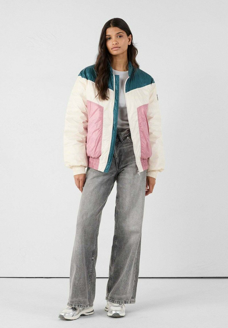 Donna in piedi che indossa un giubbotto imbottito a pannelli colorati nei toni del teal, bianco e rosa, jeans grigi a gamba larga e sneakers argento.