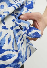 Tela de color azul y blanco con un diseño de hojas; lino texturizado; pequeña etiqueta negra visible en la costura, sostenida por una mano cerca de un bolsillo.