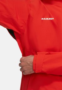 Giacca impermeabile rossa con finitura testurizzata, dotata di una tasca con zip e polsini regolabili. Logo "MAMMUT" visibile sul petto.